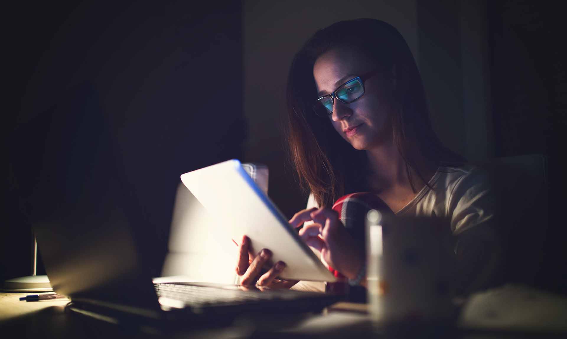 Young woman working late in front of a monitor. Businesswoman drinking coffee at her work place at night. Beautiful lady is surfing the internet late. Woman using tablet